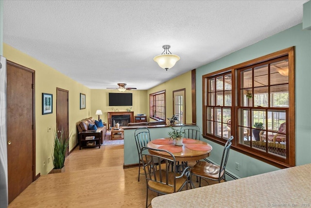 dining room featuring a fireplace, baseboards, a textured ceiling, a ceiling fan, and light wood-style flooring
