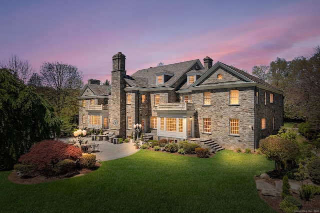 rear view of house with a lawn, a chimney, a balcony, and stone siding