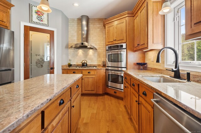 kitchen featuring wall chimney range hood, appliances with stainless steel finishes, a sink, light wood-type flooring, and decorative backsplash