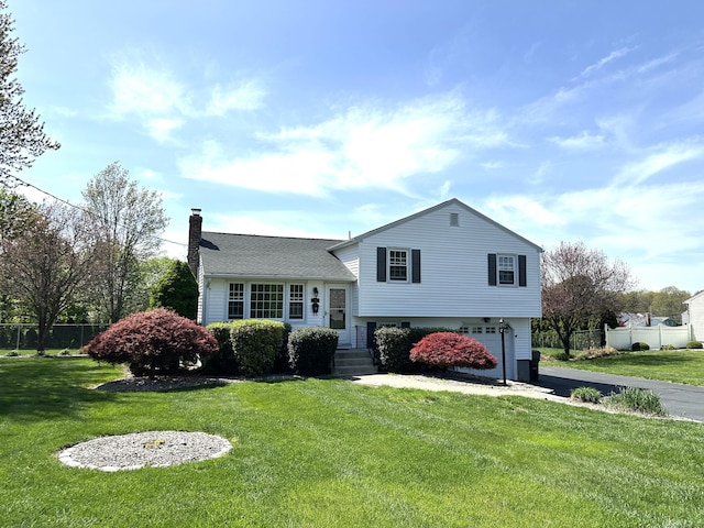 tri-level home featuring aphalt driveway, fence, a front yard, a chimney, and an attached garage