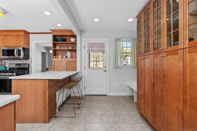 kitchen featuring brown cabinetry, appliances with stainless steel finishes, baseboards, a peninsula, and glass insert cabinets