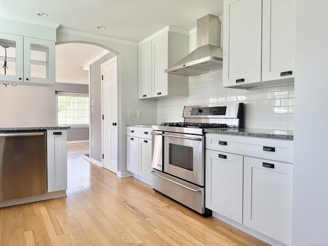 kitchen featuring arched walkways, stone countertops, wall chimney exhaust hood, stainless steel appliances, and white cabinetry