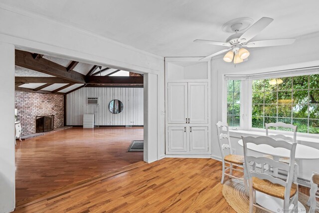 dining space featuring a ceiling fan, light wood finished floors, a brick fireplace, and beamed ceiling