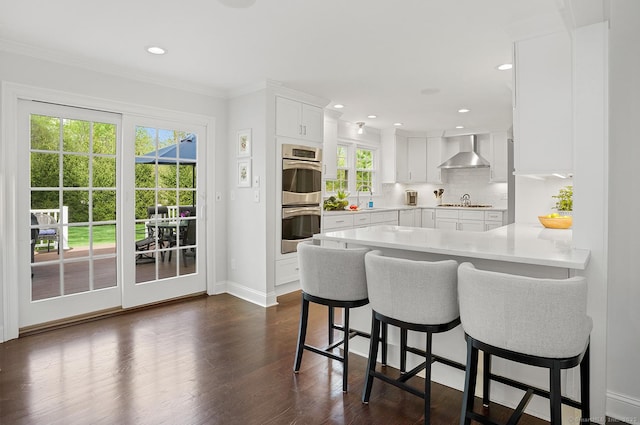 kitchen featuring double oven, a breakfast bar area, wall chimney range hood, light countertops, and white cabinetry