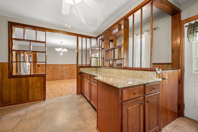 kitchen with wooden walls, brown cabinets, light stone counters, ceiling fan with notable chandelier, and a wainscoted wall