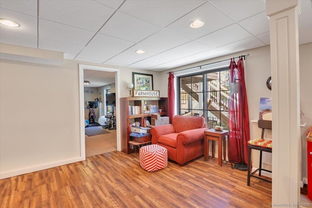 living area with baseboards, decorative columns, a drop ceiling, and wood finished floors