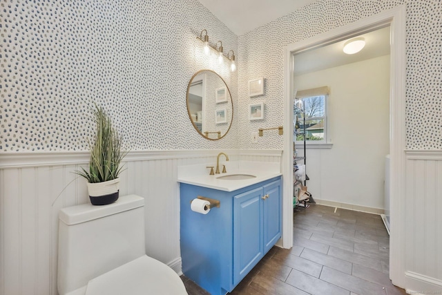 bathroom with vanity, wainscoting, toilet, and wallpapered walls