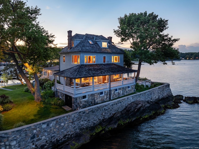 back of house featuring stone siding, a water view, a chimney, a balcony, and a lawn
