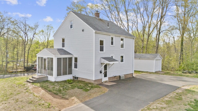 view of side of property featuring an outdoor structure, a chimney, a sunroom, a shingled roof, and driveway