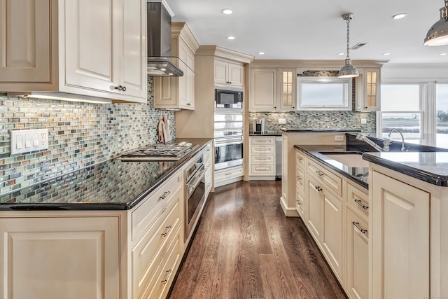 kitchen featuring cream cabinets, appliances with stainless steel finishes, wall chimney exhaust hood, tasteful backsplash, and dark wood-style floors