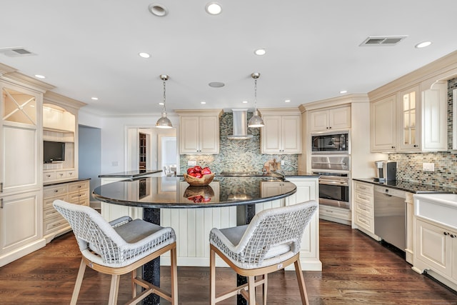 kitchen with visible vents, dark countertops, wall chimney exhaust hood, cream cabinetry, and stainless steel appliances