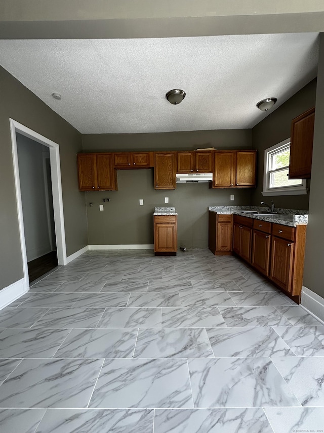 kitchen with brown cabinetry, marble finish floor, baseboards, a textured ceiling, and under cabinet range hood