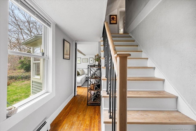 staircase with a textured ceiling, a baseboard heating unit, and wood finished floors