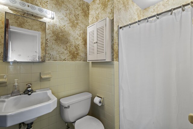 bathroom featuring tile walls, wainscoting, a sink, toilet, and wallpapered walls