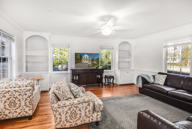 living room with built in shelves, crown molding, wood finished floors, a ceiling fan, and a wainscoted wall