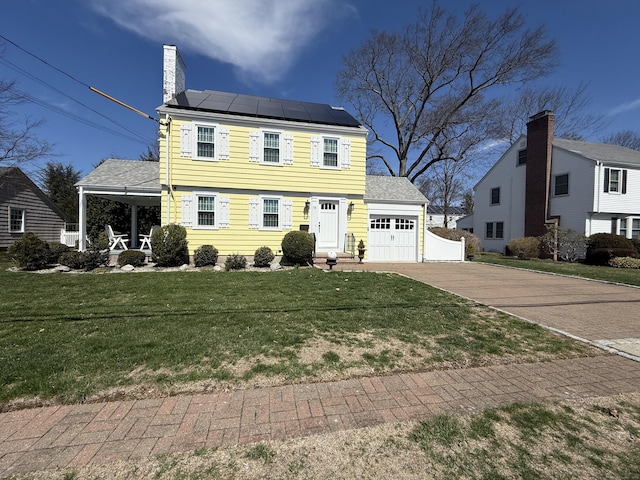 colonial home featuring driveway, a chimney, roof mounted solar panels, a front lawn, and an attached garage