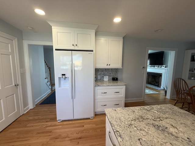 kitchen featuring backsplash, white refrigerator with ice dispenser, a brick fireplace, light wood-type flooring, and white cabinetry