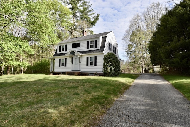 colonial inspired home with aphalt driveway, a front yard, a gambrel roof, roof with shingles, and a chimney