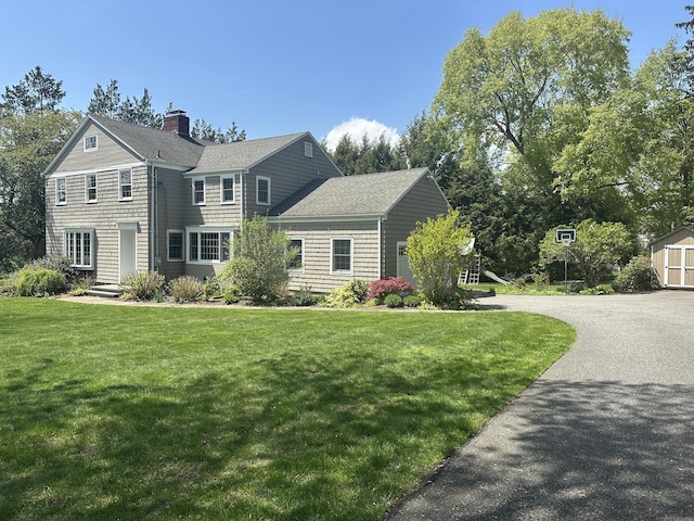 view of front facade with a shingled roof, a chimney, aphalt driveway, and a front yard