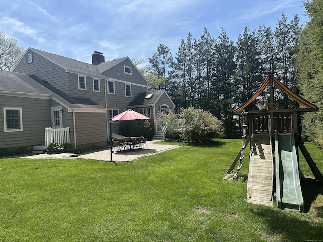 rear view of house with a chimney, a shingled roof, a patio area, a playground, and a lawn