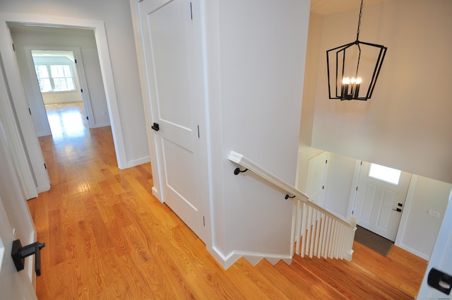 hall with light wood-style floors, baseboards, a chandelier, and an upstairs landing