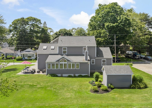 back of property with a lawn, a patio, an outbuilding, and a shingled roof