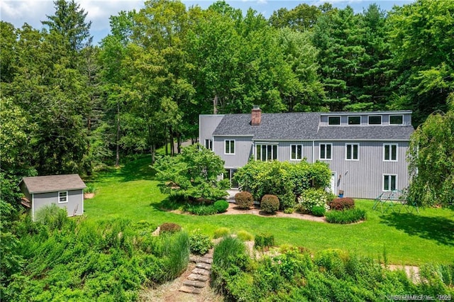 view of front of home featuring a wooded view, a chimney, a shingled roof, an outdoor structure, and a front yard