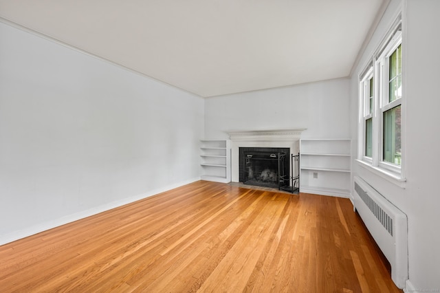 unfurnished living room with radiator, built in features, baseboards, a fireplace with flush hearth, and light wood-style flooring