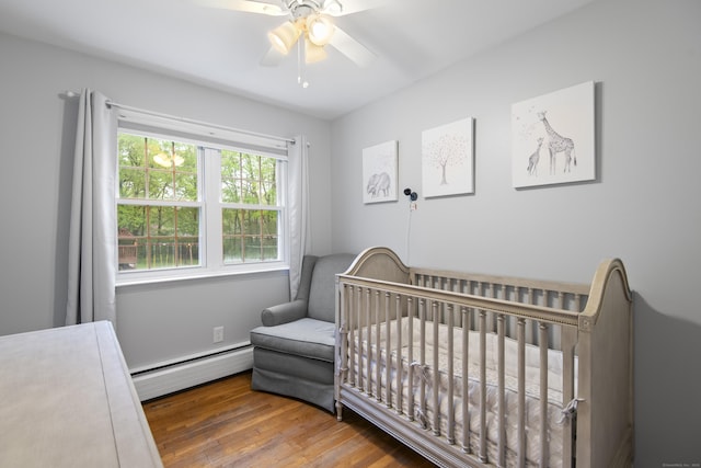 bedroom featuring baseboard heating, wood finished floors, a nursery area, and a ceiling fan