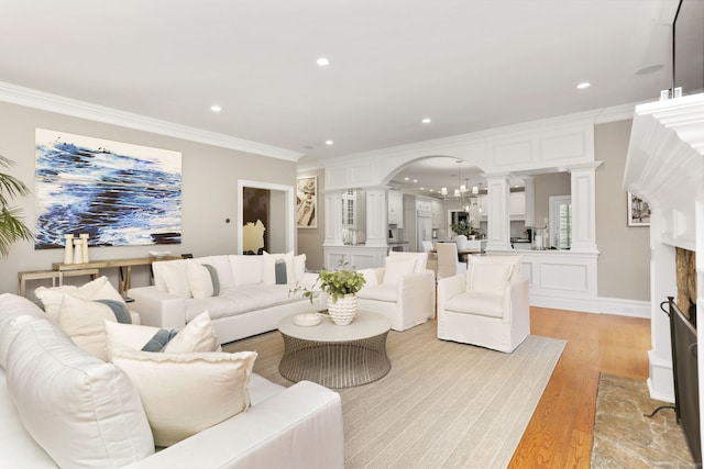 living area featuring light wood-style flooring, ornate columns, crown molding, arched walkways, and recessed lighting