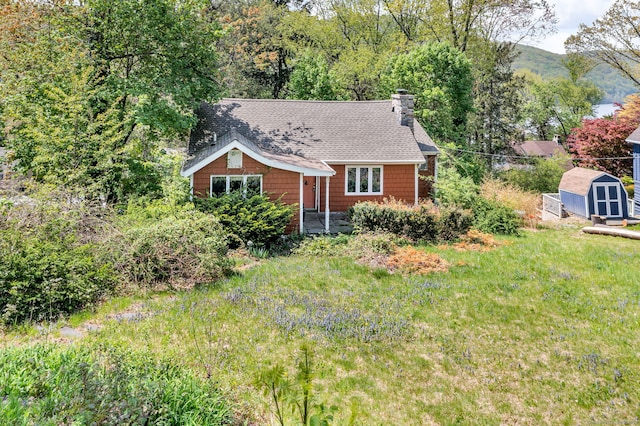 view of front facade featuring a chimney, a shed, an outdoor structure, roof with shingles, and a front lawn