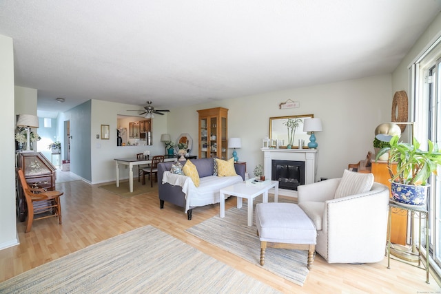 living room with light wood-type flooring, baseboards, a ceiling fan, and a glass covered fireplace
