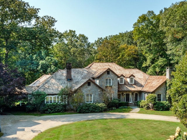 view of front facade with stone siding, a chimney, a front yard, curved driveway, and a high end roof