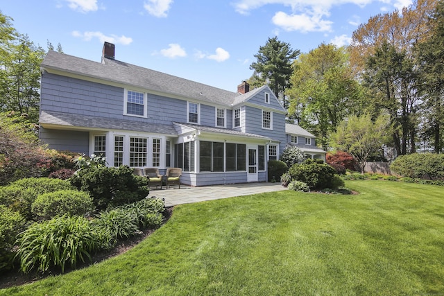 rear view of property with a chimney, roof with shingles, a sunroom, a yard, and a patio
