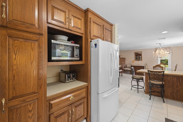 kitchen featuring light tile patterned floors, freestanding refrigerator, brown cabinetry, stainless steel microwave, and decorative light fixtures