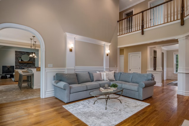 living room featuring ornate columns, a decorative wall, a towering ceiling, wood finished floors, and a wainscoted wall
