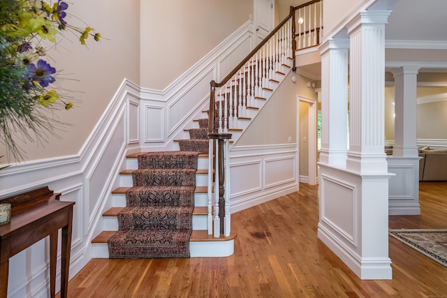 staircase featuring a decorative wall, wood finished floors, crown molding, ornate columns, and wainscoting