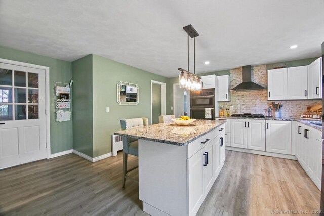 kitchen with wall chimney exhaust hood, multiple ovens, light wood finished floors, white cabinetry, and recessed lighting