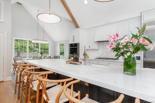 kitchen featuring beam ceiling, light wood-style flooring, high vaulted ceiling, light stone counters, and appliances with stainless steel finishes