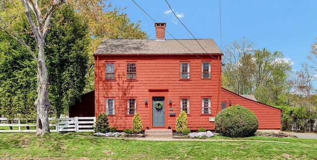colonial home with roof with shingles, a chimney, fence, and a front yard