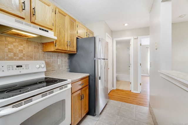 kitchen featuring freestanding refrigerator, under cabinet range hood, light tile patterned floors, white range with electric stovetop, and backsplash