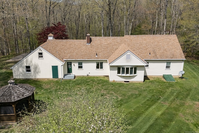 rear view of property featuring a lawn, a chimney, a wooded view, and a shingled roof