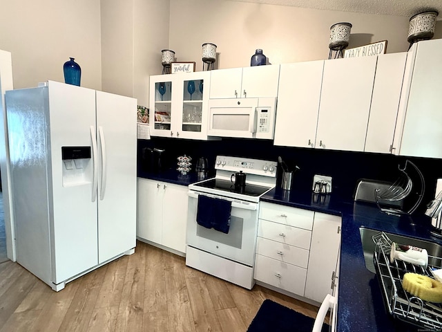 kitchen featuring glass insert cabinets, white appliances, light wood-style floors, white cabinetry, and dark countertops