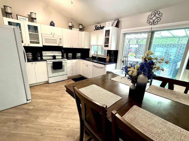 kitchen with a sink, white appliances, light wood-type flooring, vaulted ceiling, and dark countertops