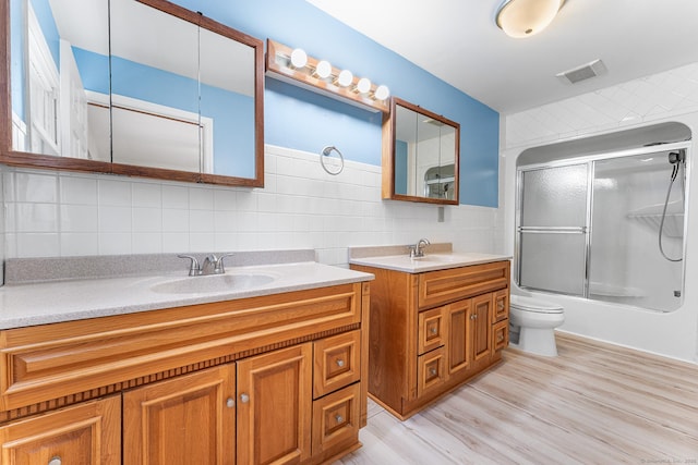 full bath featuring backsplash, two vanities, bath / shower combo with glass door, toilet, and wood finished floors