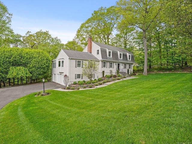 dutch colonial featuring driveway, a garage, a chimney, a shingled roof, and a front lawn