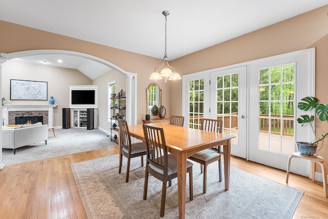 dining area featuring ornate columns, arched walkways, light wood-type flooring, a lit fireplace, and vaulted ceiling
