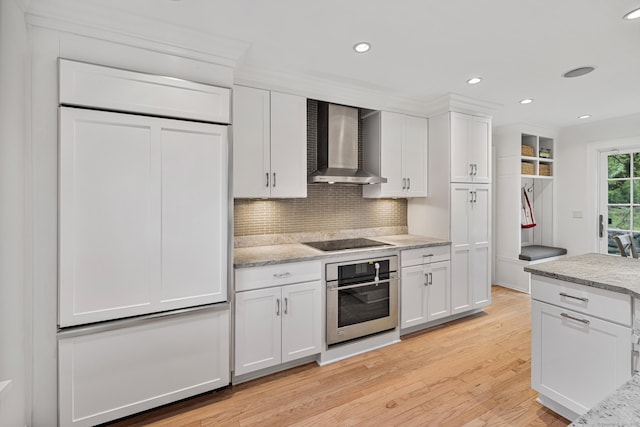 kitchen featuring stainless steel oven, wall chimney range hood, black electric cooktop, light wood finished floors, and white cabinetry