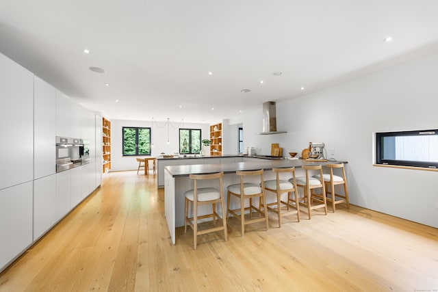 kitchen featuring wall chimney range hood, modern cabinets, white cabinetry, light wood-type flooring, and a kitchen bar