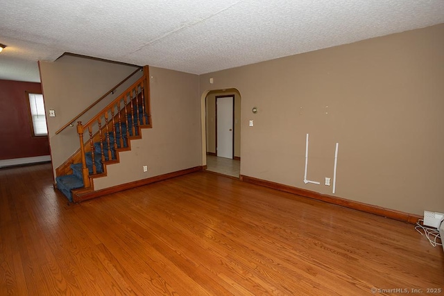 unfurnished living room with arched walkways, light wood-style flooring, stairway, a textured ceiling, and baseboards